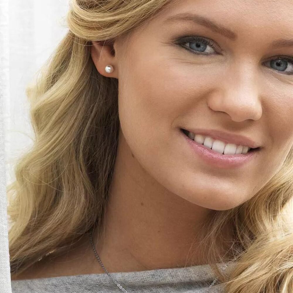 Close-up of a smiling woman with blonde hair and pearl earrings, showcasing a natural look and clear skin.