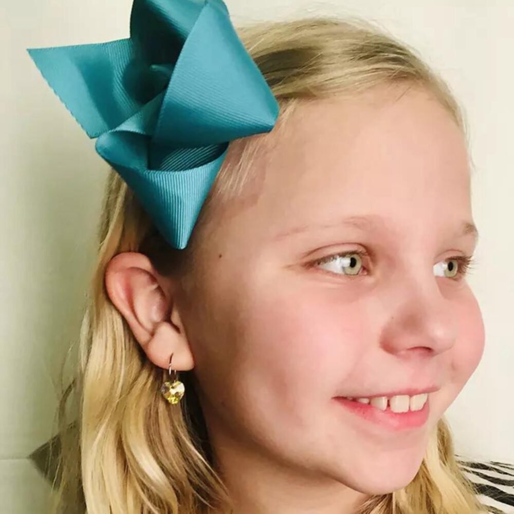 A young girl smiling with a turquoise hair bow and yellow earrings, showcasing a cheerful expression and light blonde hair.