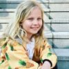 A young girl with blonde hair wearing a pineapple-patterned jacket, sitting on stone steps, smiling brightly with blue eyes.