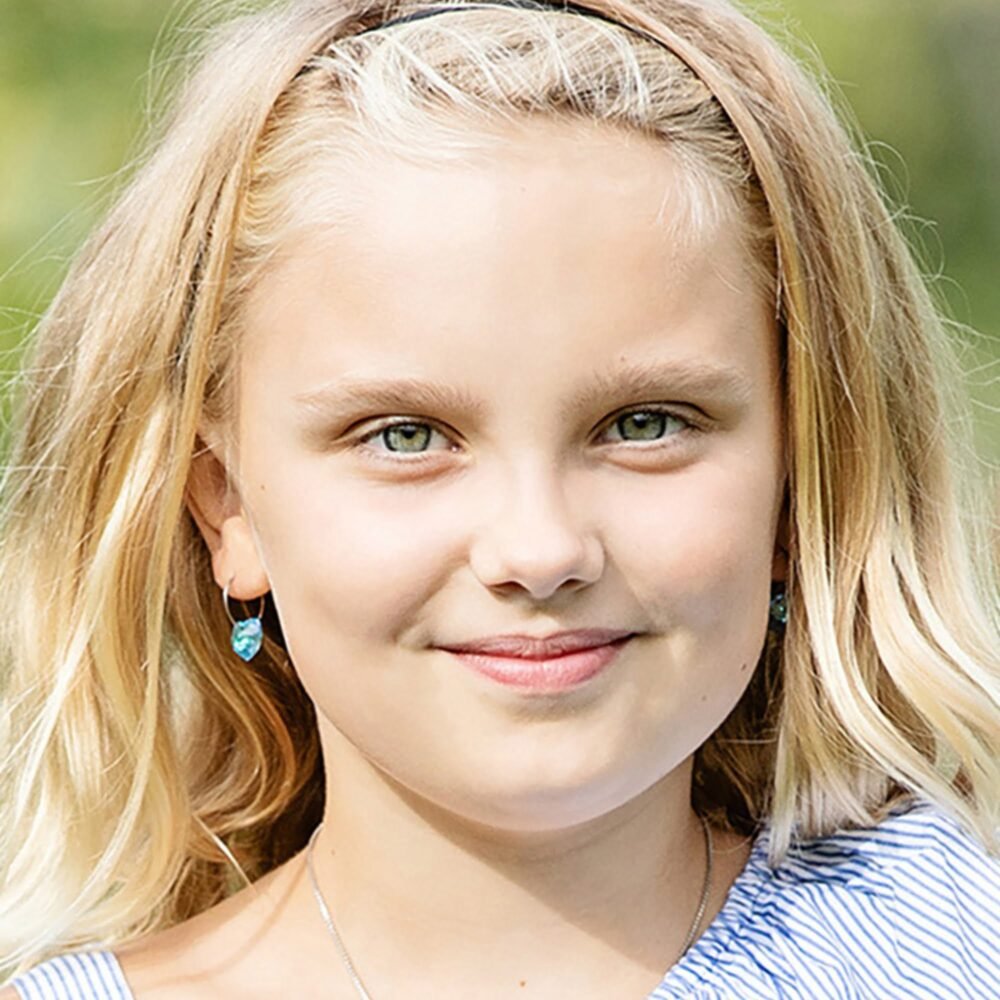 A young girl with blonde hair and blue earrings, smiling at the camera, showcasing her bright green eyes and cheerful expression.