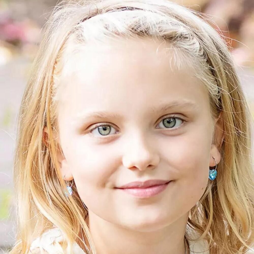 A young girl with blonde hair and blue earrings, smiling at the camera, showcasing her bright blue eyes and cheerful expression.