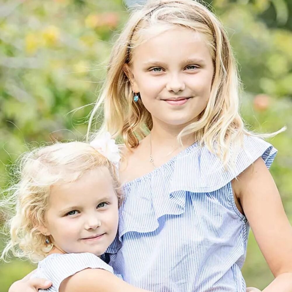 Two young girls wearing matching striped outfits, one with blonde hair and blue eyes, the other with curly blonde hair, smiling outdoors.