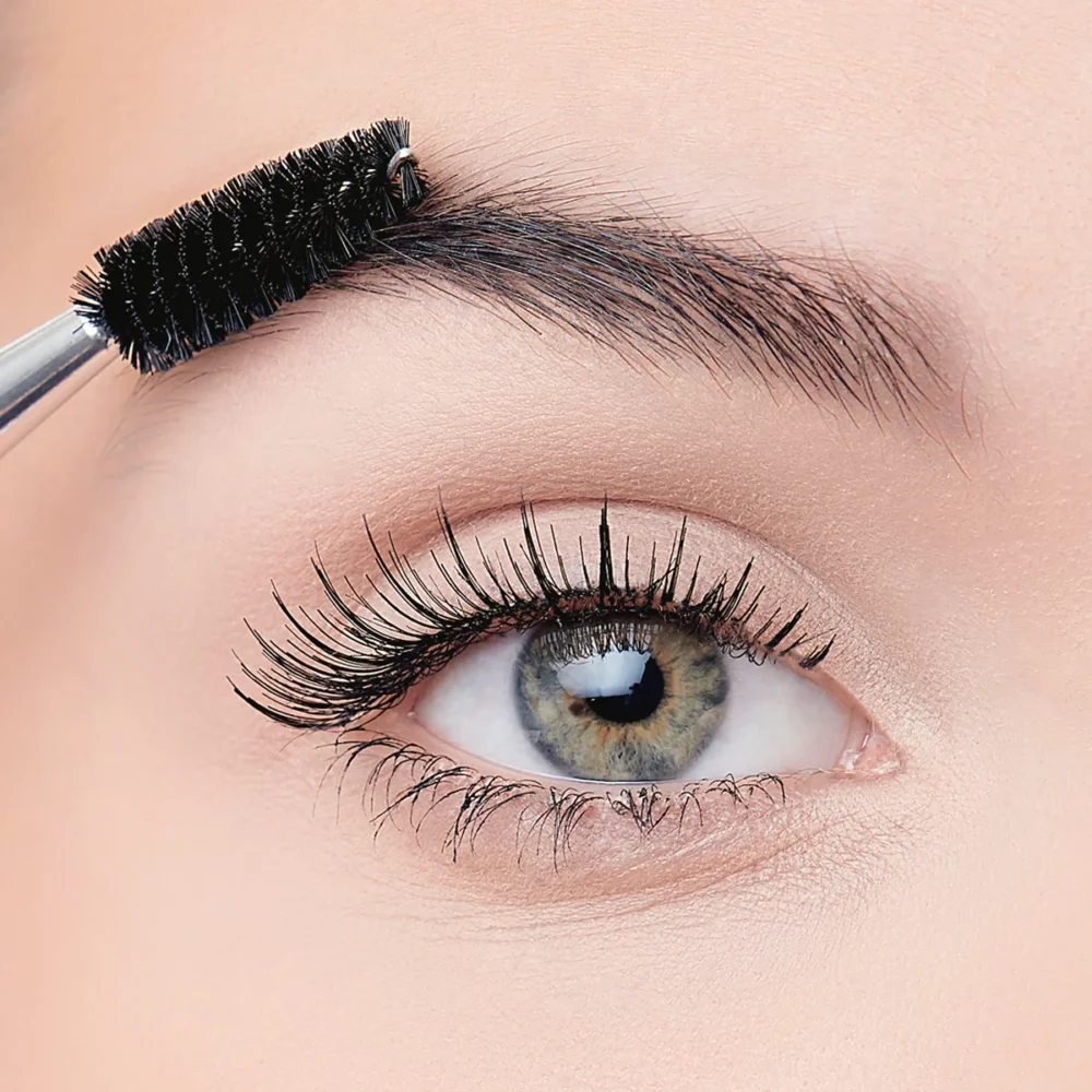 Close-up of a woman's eye with long eyelashes and an eyebrow brush grooming the brows, showcasing beauty and makeup application.