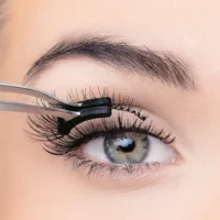 Close-up image of a person applying false eyelashes using tweezers, showcasing long, voluminous lashes and a detailed eye.