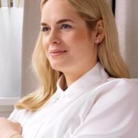 Profile of a woman with blonde hair, wearing a white blouse and small earrings, smiling softly in a well-lit indoor setting.