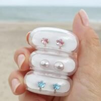 Three pairs of earrings in a clear case, featuring pink flowers, blue flowers, and pearl studs, ideal for jewelry enthusiasts.