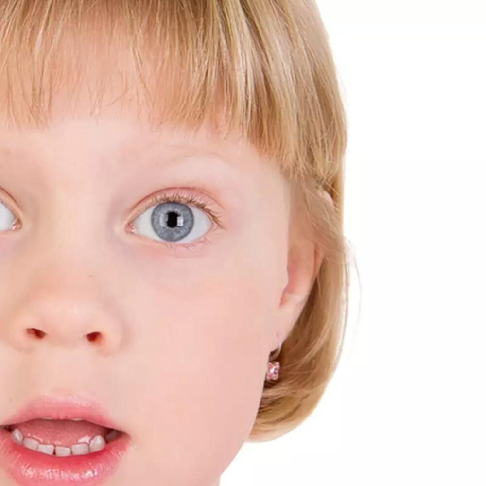 A surprised young girl with short blonde hair and blue eyes, wearing a pink earring, expressing shock or amazement.