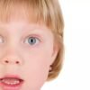 A surprised young girl with short blonde hair and blue eyes, wearing a pink earring, expressing shock or amazement.