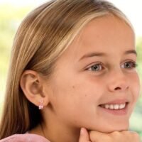 A young girl with light brown hair smiling, showcasing pink bow earrings. The earrings feature a small gemstone accent, adding charm to her look.