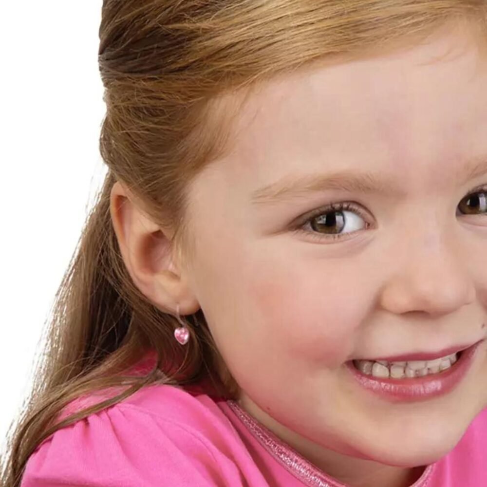 A young girl with long brown hair smiles while wearing pink clothing and heart-shaped earrings, showcasing a cheerful expression.