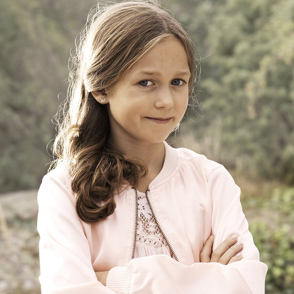 A young girl with long brown hair wearing a pink bomber jacket, standing confidently with arms crossed in a natural setting.