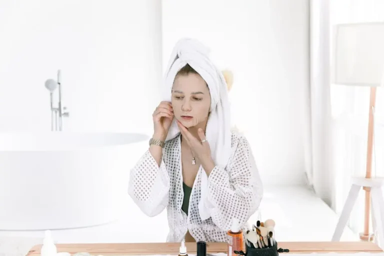 A woman in a white robe and towel applying skincare products at a vanity with various beauty items, showcasing a skincare routine.